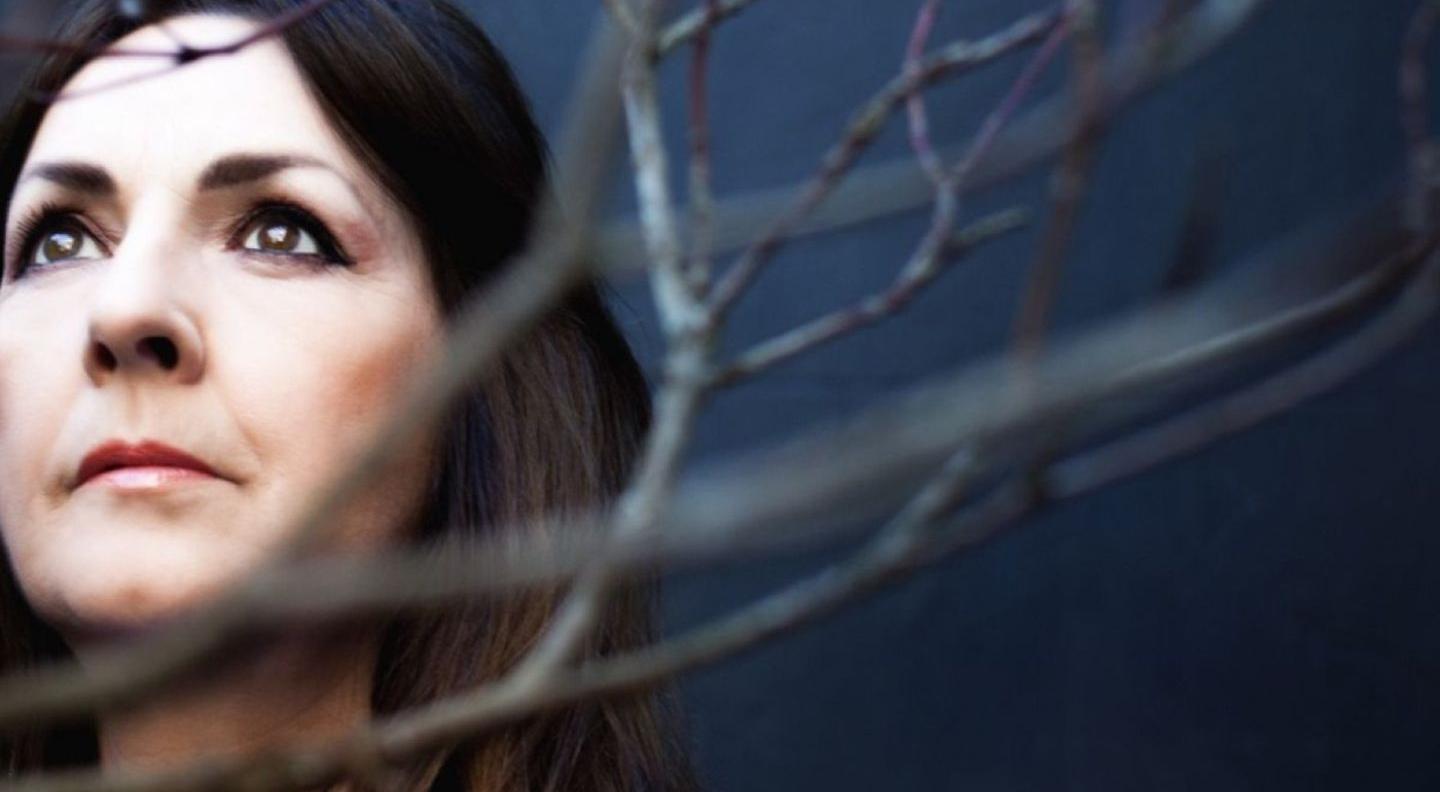 A white woman with dark hair and a blue shirt stands among the bare branches of a tree, looking upwards