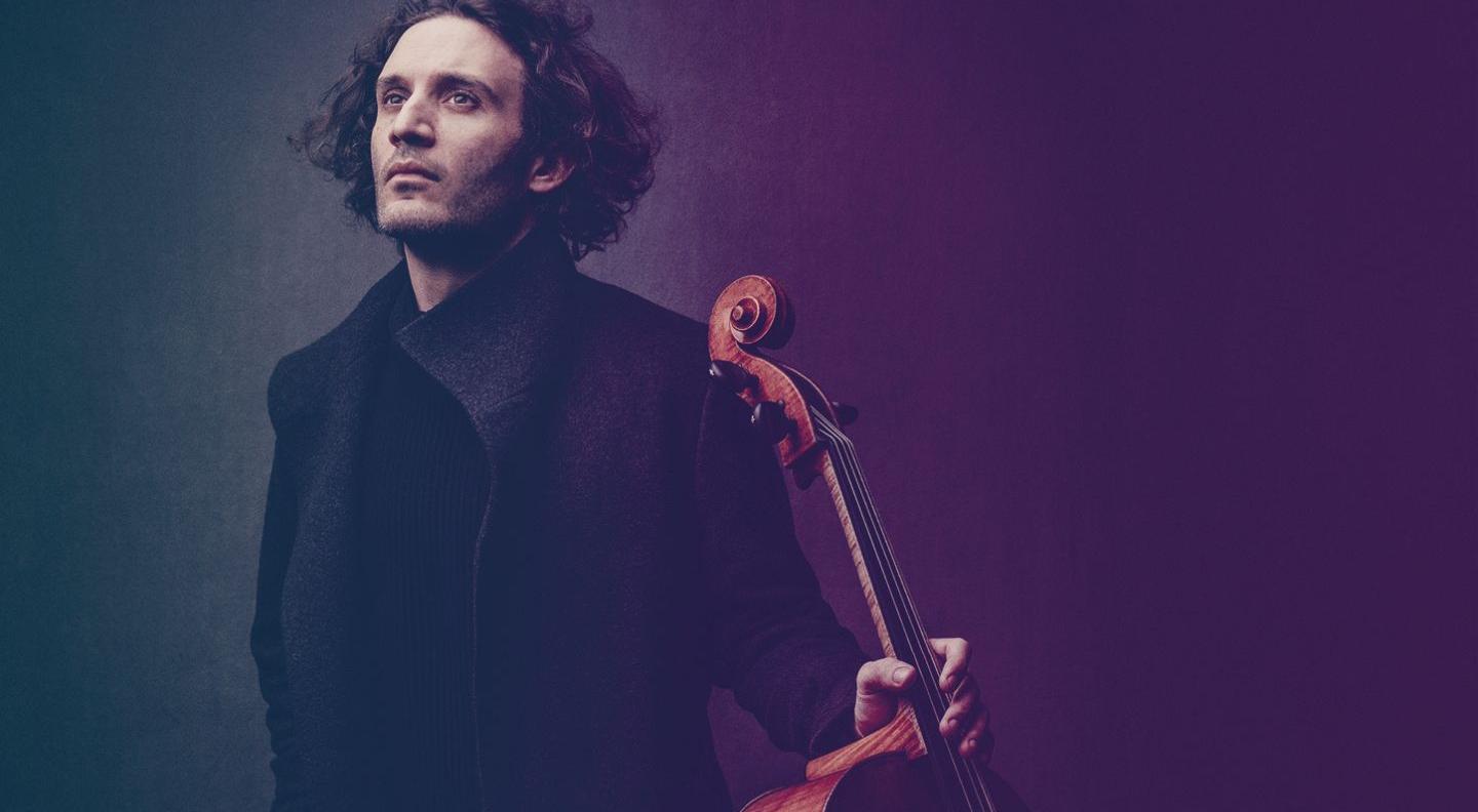 A young man with long curly dark hair, wearing a dark wool coat. He holds the neck of a cello