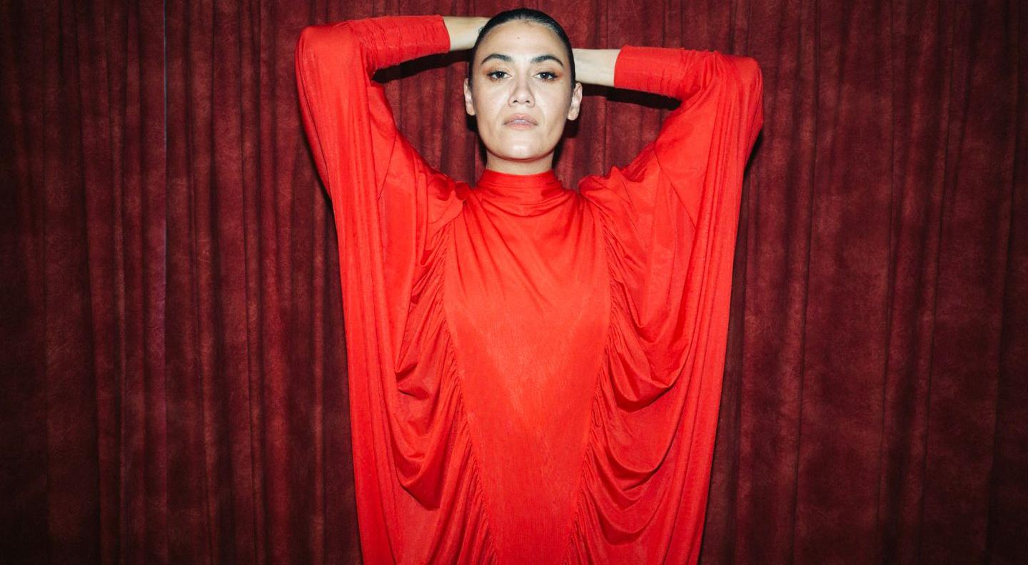 an image of Nadine Shah against a dark red background, wearing a bright red dress. She has her hands on her head, and stares into the camera