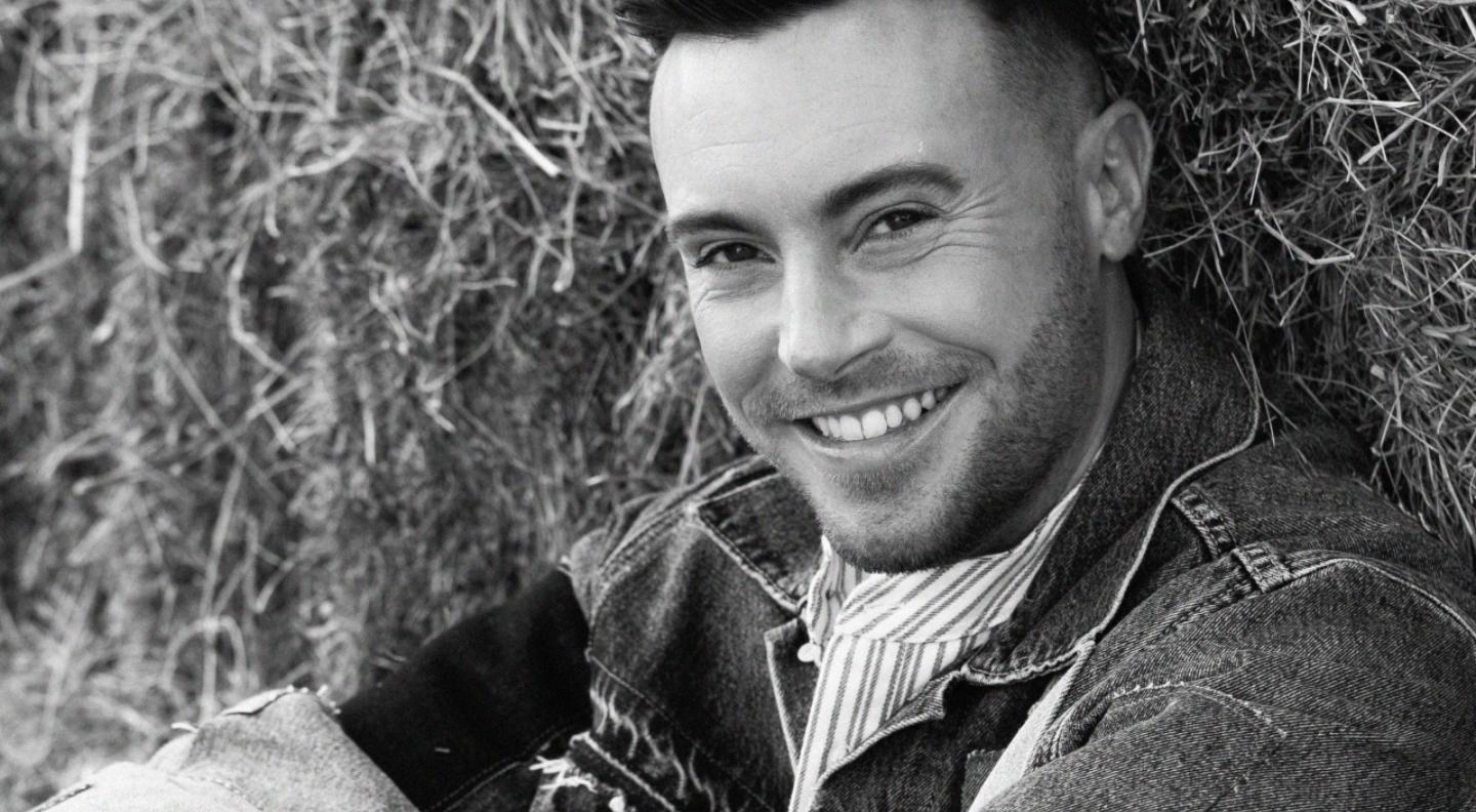 A black and white midshot of Nathan sitting against a hay bale smiling at the camera