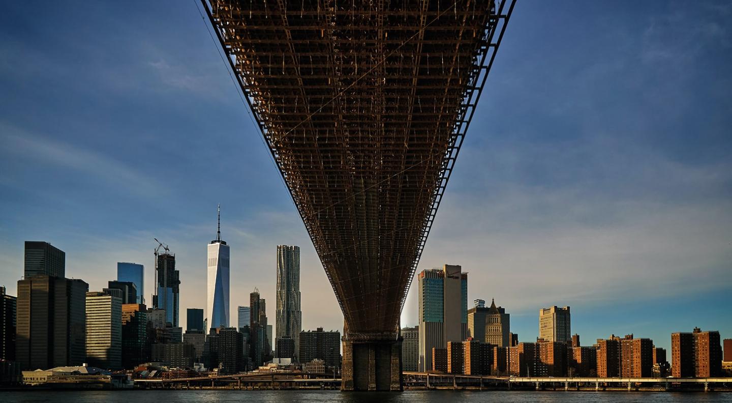 A wide shot of the view of the skyscrapers of Manhattan from under one of the bridges spanning the Hudson or East River.