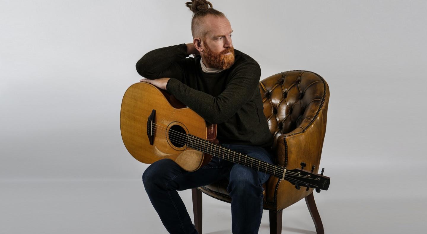 Newton Faulkner sits on a leather chair, with an acoustic guitar in hand
