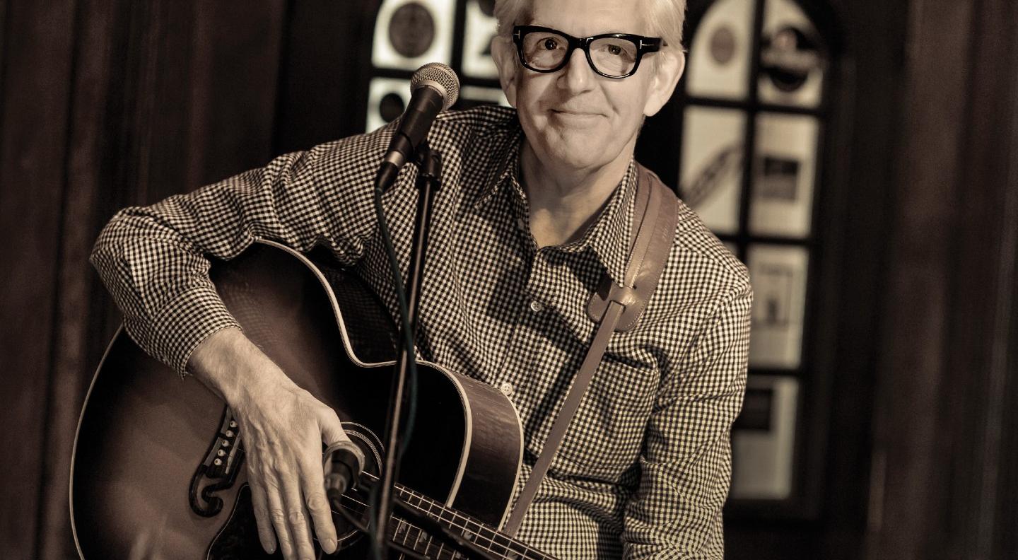 A black and white mid shot of Nick Lowe seated at a microphone with his guitar on his knee smiling at the camera