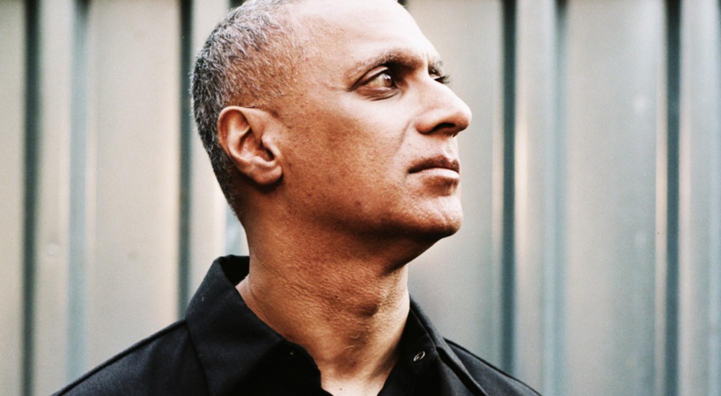 An image of Nitin Sawhney's head and shoulders. He wears a dark shirt and stands in front of what looks like a shipping container, looking off to the right of the image