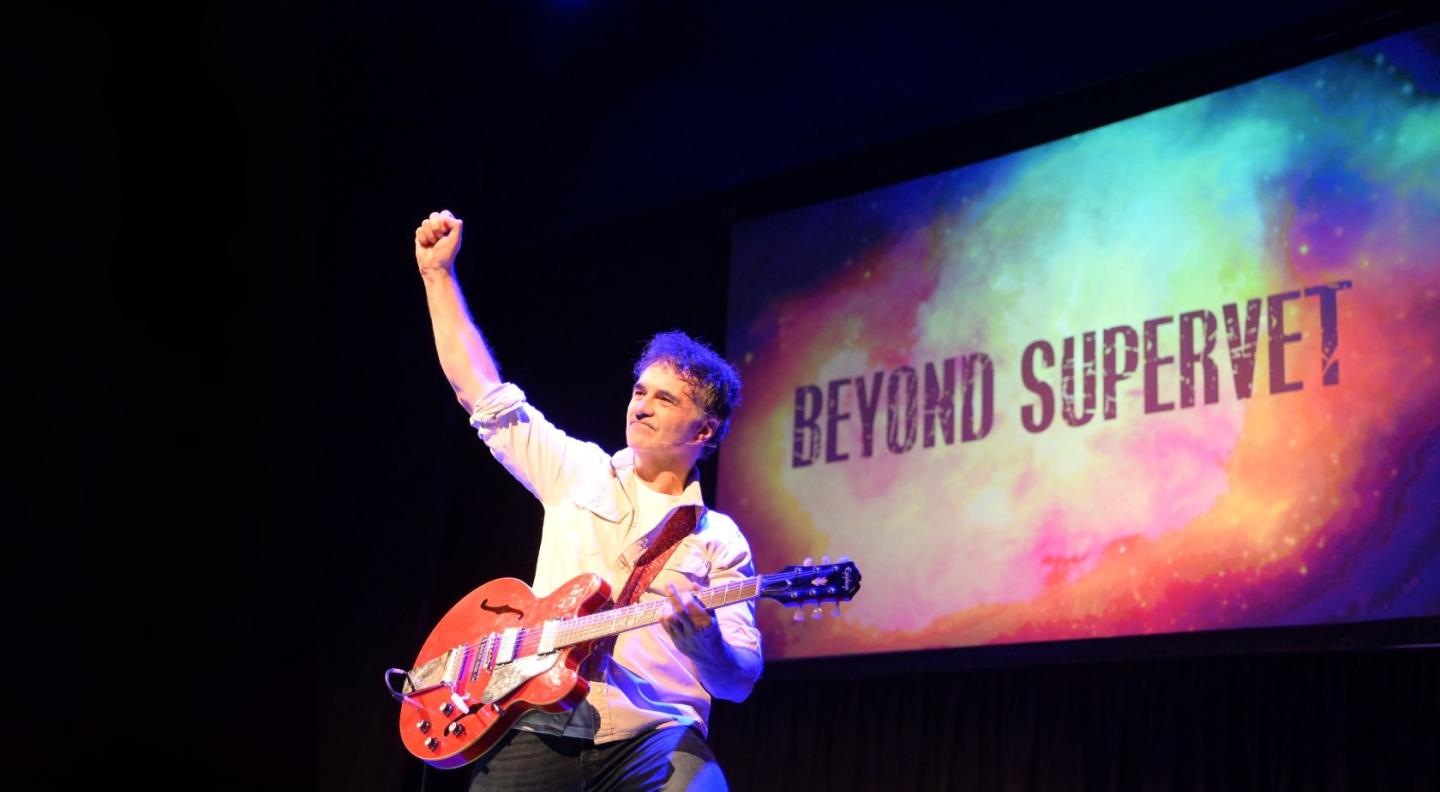 Noel Fitzpatrick with a guitar in front of a screen that says 'beyond supervet'