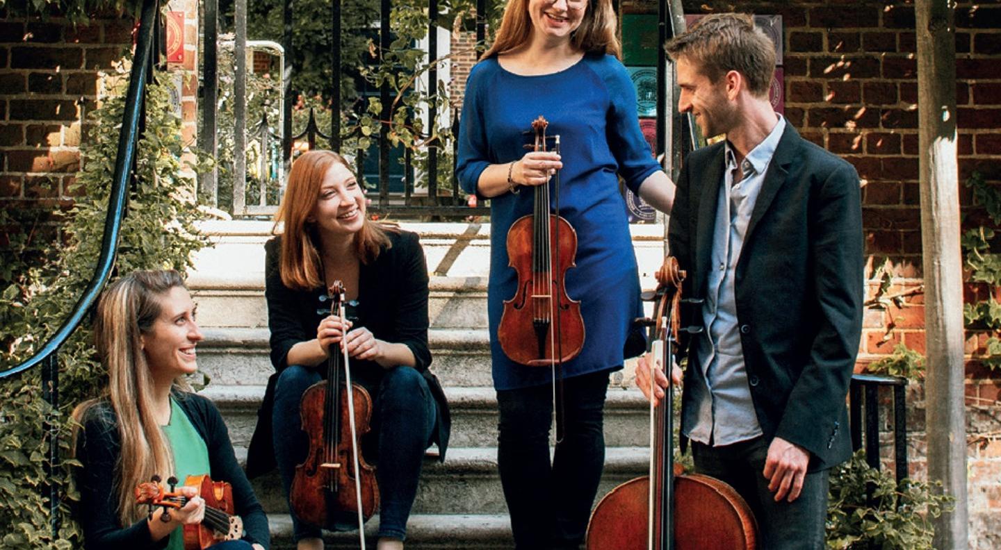 A full length shot of the quartet sitting and standing in what looks like a garden setting with steps and wrought iron railings. They all smile and hold their instruments