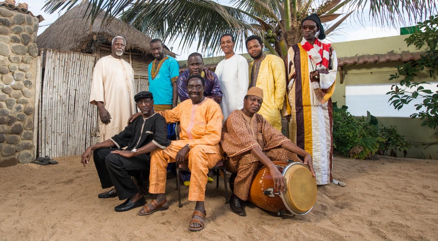 The various members of Orchestra Baobab sit and stand in a group in an African setting, with sand, palm trees and thatchd buildings surrounding them