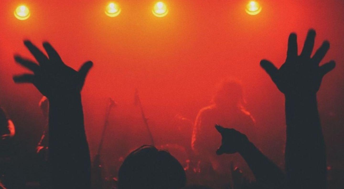 The back of a crowd, looking towards a stage, which is lit in red and very hazy. There are silhouettes of heads and the backs of arms held up