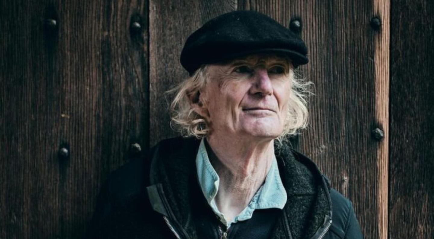 Peter Hammill, wearing a black flat cap, standing in front of a wood backround.