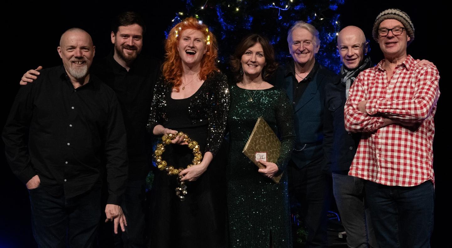 The line-up for Phil's Christmas Songbook standing in a line smiling and  laughing at the camera in front of a festive background