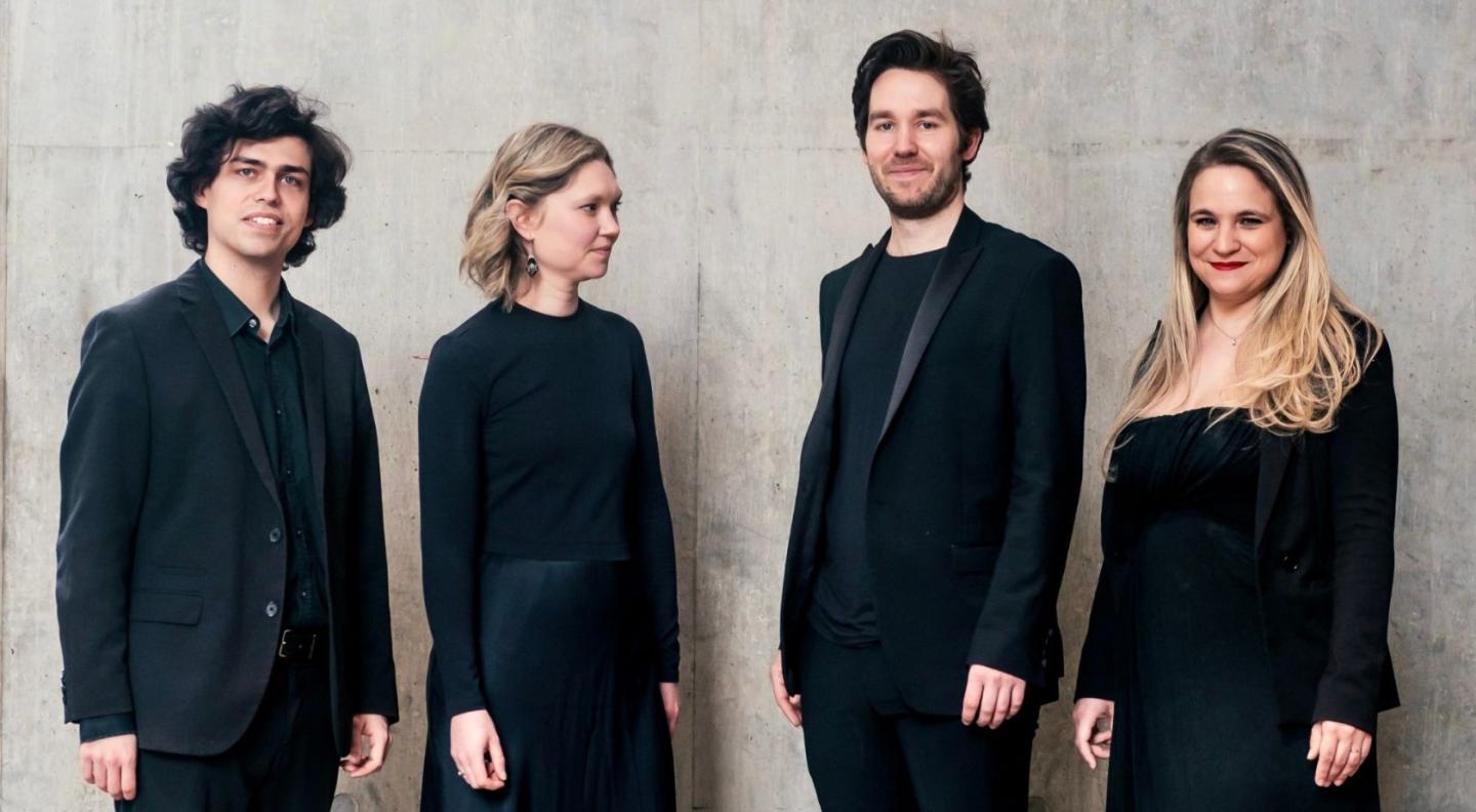 The four members of the Piatti Quartet, two women and two men, standing in a line, all dressed in concert black clothing.