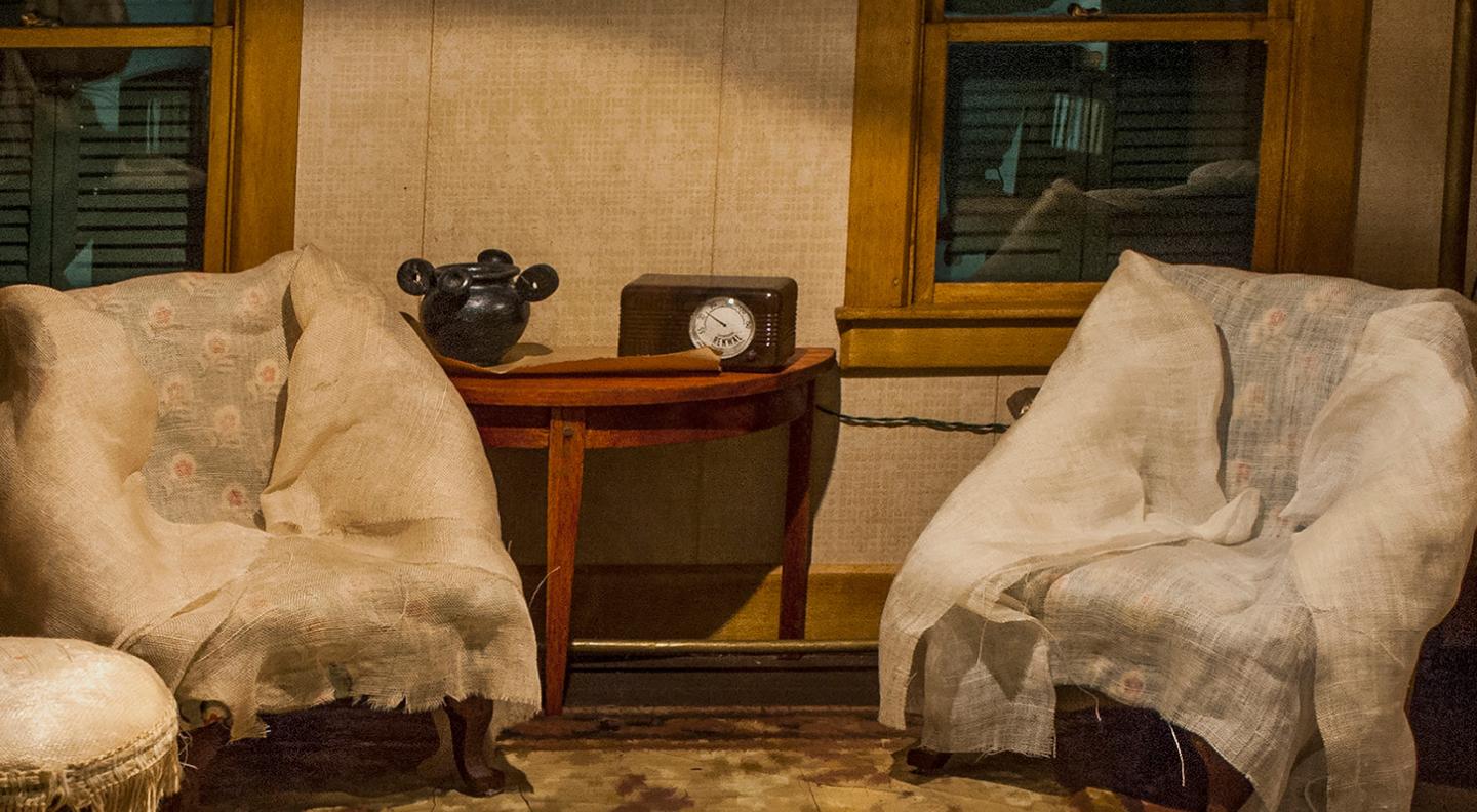 An image of what looks like the stage set of a period front room. Two armchairs covered in white cloth are either side of a table with a black vase on and old-fashioned wireless radio on it. Two closed and shuttered windows are behind the chairs 