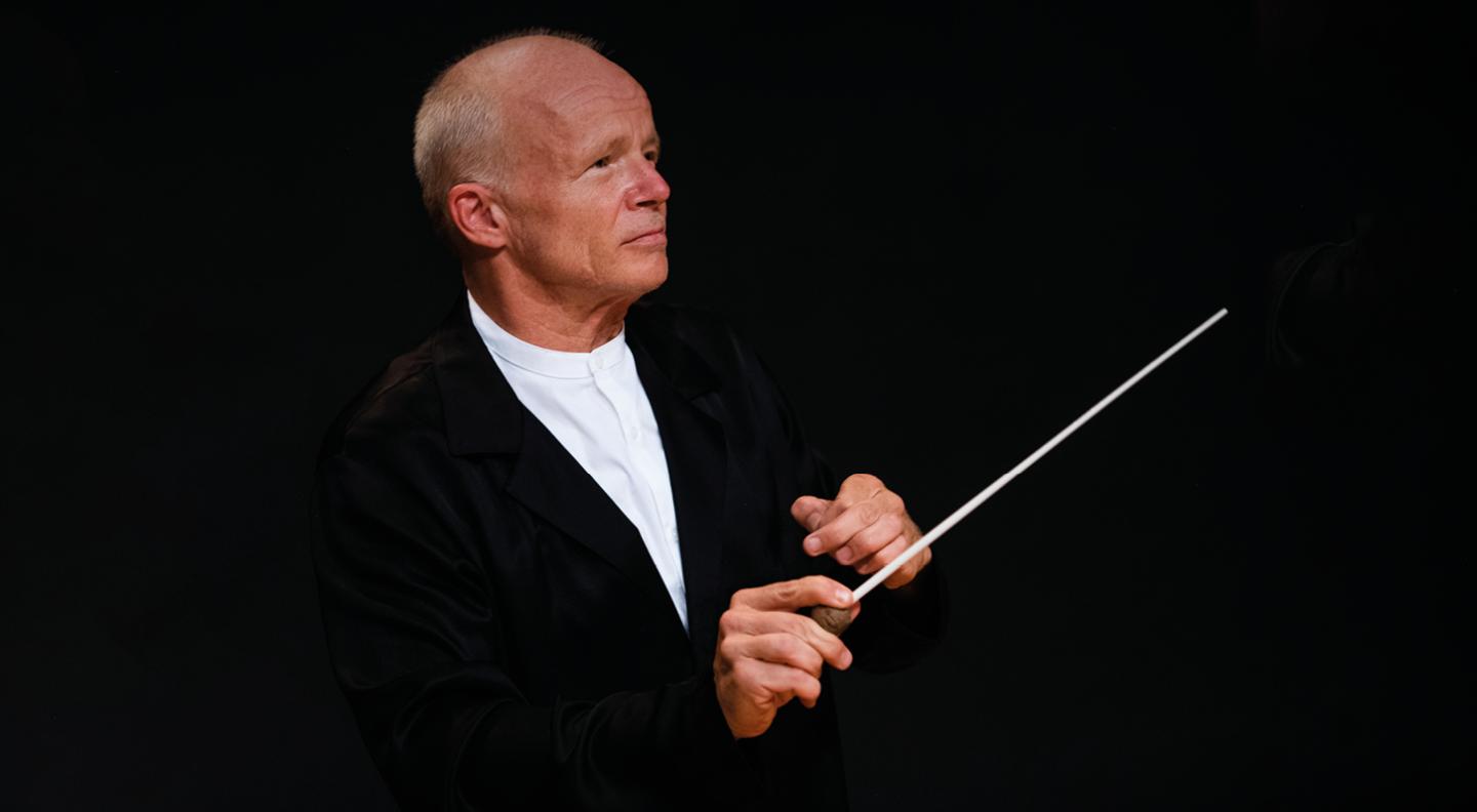 A mid shot of Thomas Zehetmair in a black jacket and white shirt against a black background, holding a baton ready to conduct