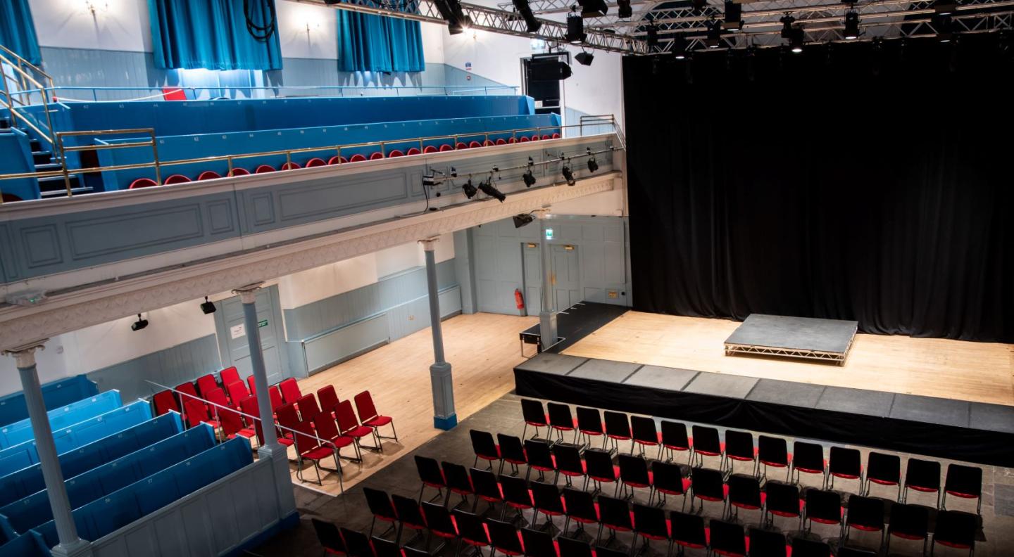 A view from the gallery across to The Queen's Hall stage showing red stall seats and blue pew seating