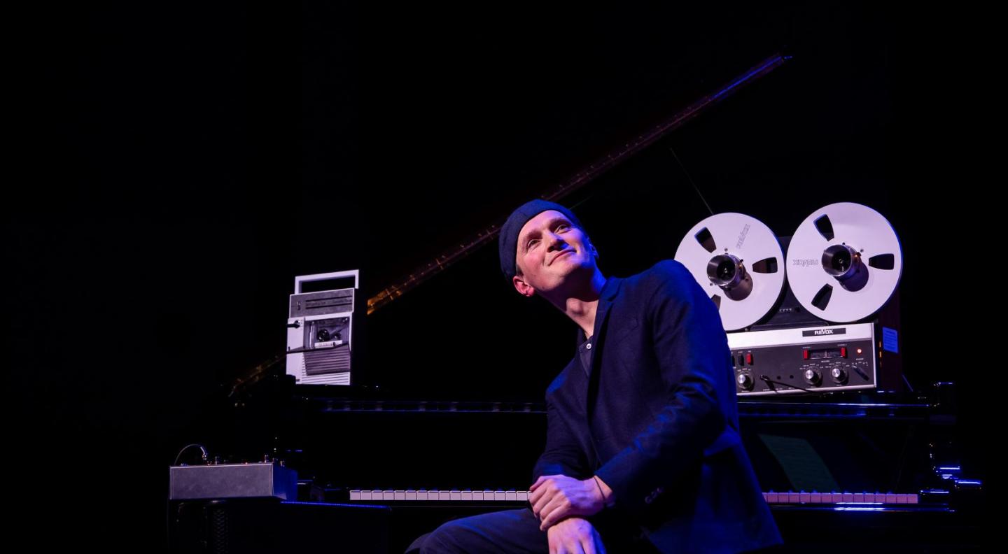 A white man in dark suit wearing a dark cap backwards sits on a piano stool looking upwards smiling with a tape deck next to him