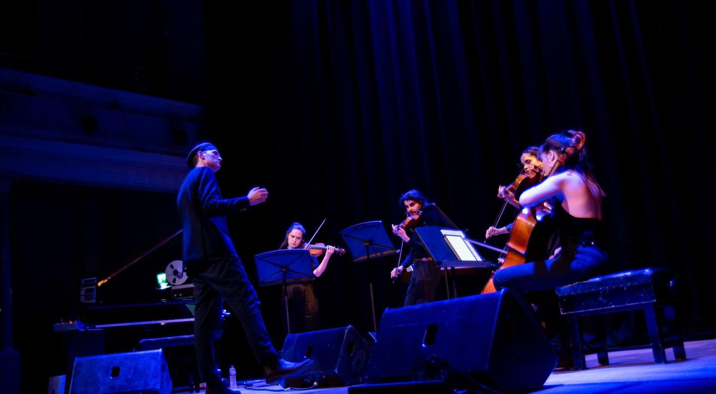 A tall white man wearing a dark suit and hat conducts a small ensemble of stringed instrument players on a dark stage