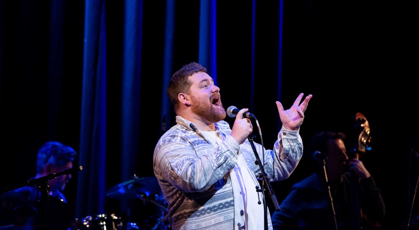 A young white man with brown hair wearing a blue shirt over a white t-shirt sings passionately into a microphone on stage