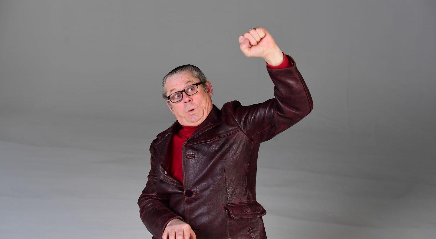 a white man wearing a red leather jacker plays a digital organ, while raising a fist in the air
