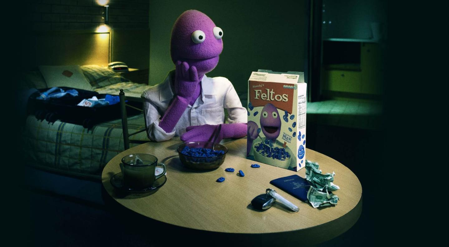 A photograph of Randy Feltface, a purple puppet, sat at a table eating a bowl of cereal called 'Felt-o's'