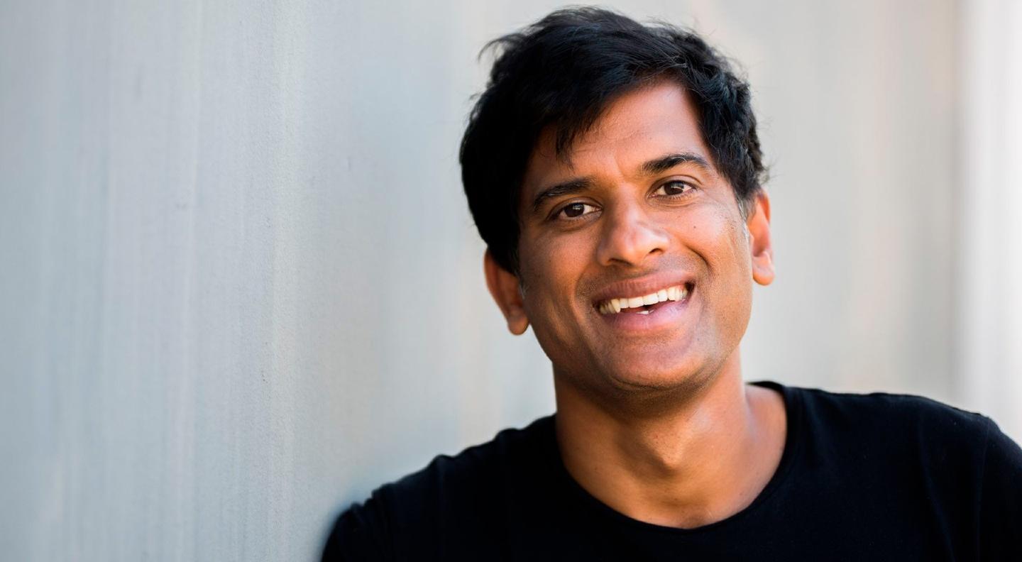 A head and shoulders shot of Rangan against a plain background. He wears a black top and smiles at the camera.