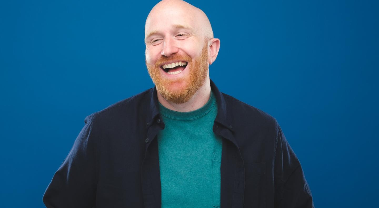 A bald white man in his thirties with a ginger beard. He wears a dark jacket and green top and is laughing