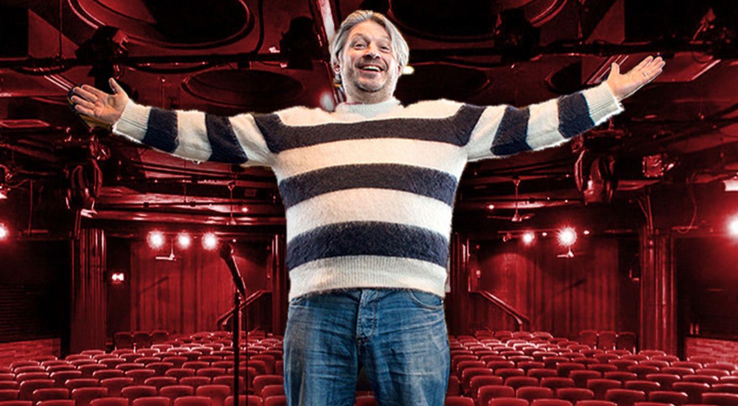 Richard Herring wears a striped jumper and  jeans, and stands on stage in front of red theatre-style seating