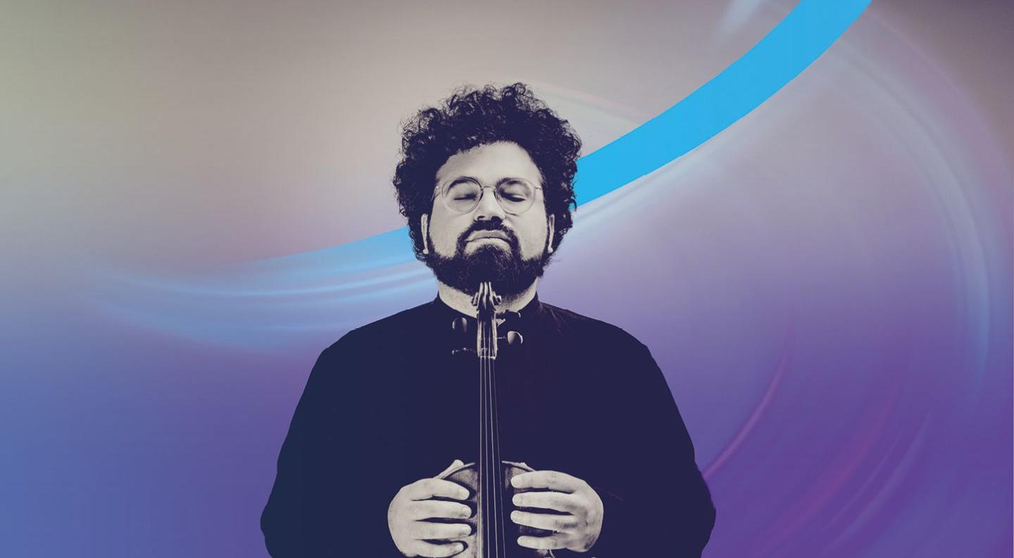 A man with curly black hair and a beard, wearing round glasses, holds a violin up to his chin