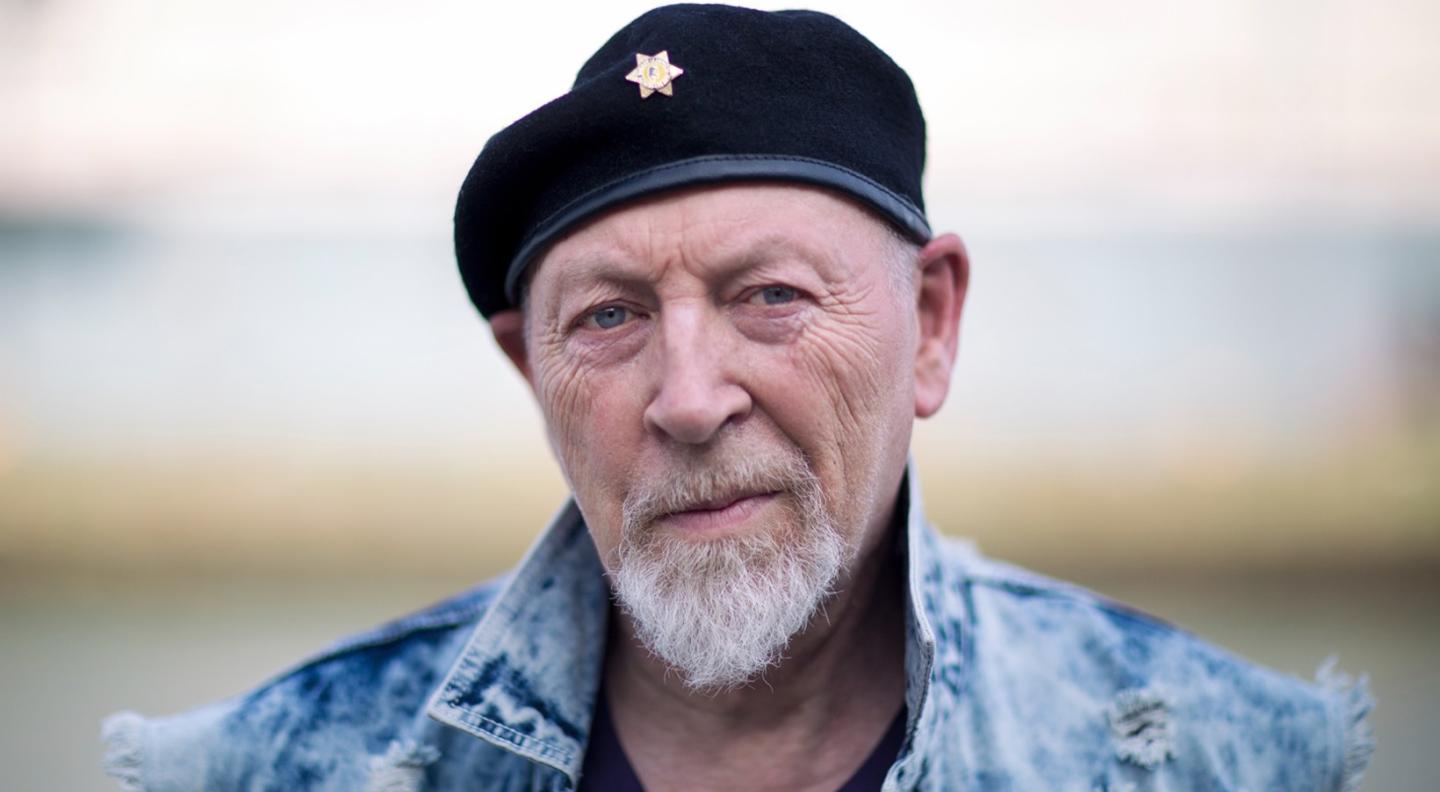 A full colour close up shot of Richard Thompson looking at the camera. He wears a black beret and a blue denim jacket