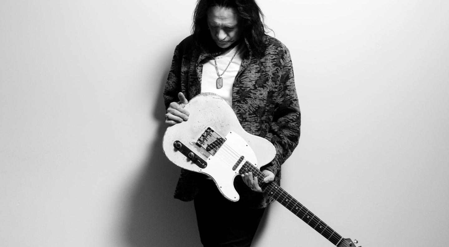 A photograph of Robben Ford holding his electric guitar from the bottom, looking down at it
