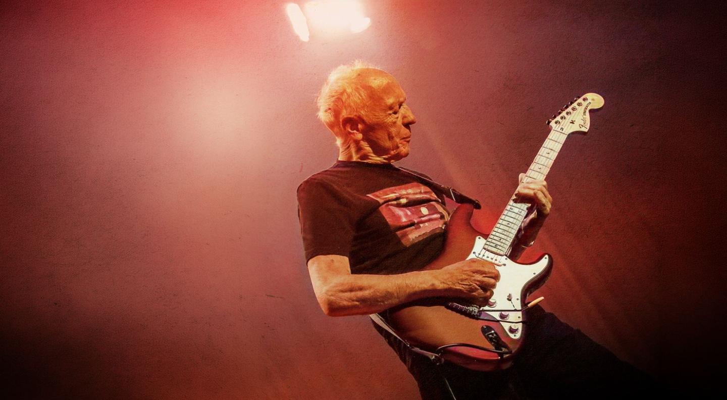 Robin Trower playing the guitar on stage with a red coloured background