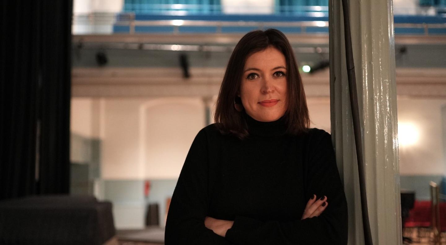 A full colour mid shot image of a white woman with shoulder length dark hair wearing a black jumper. She leans against a pillar with the lights and auditorium of The Queen's Hall visible behind her