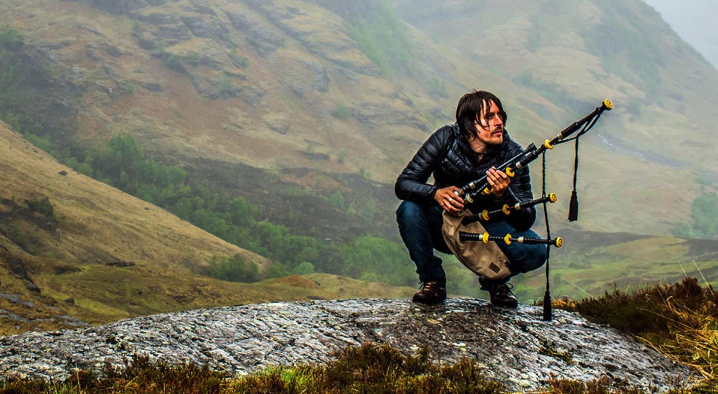 Ross Ainslie with pipes crouching against a beautiful highland backdrop