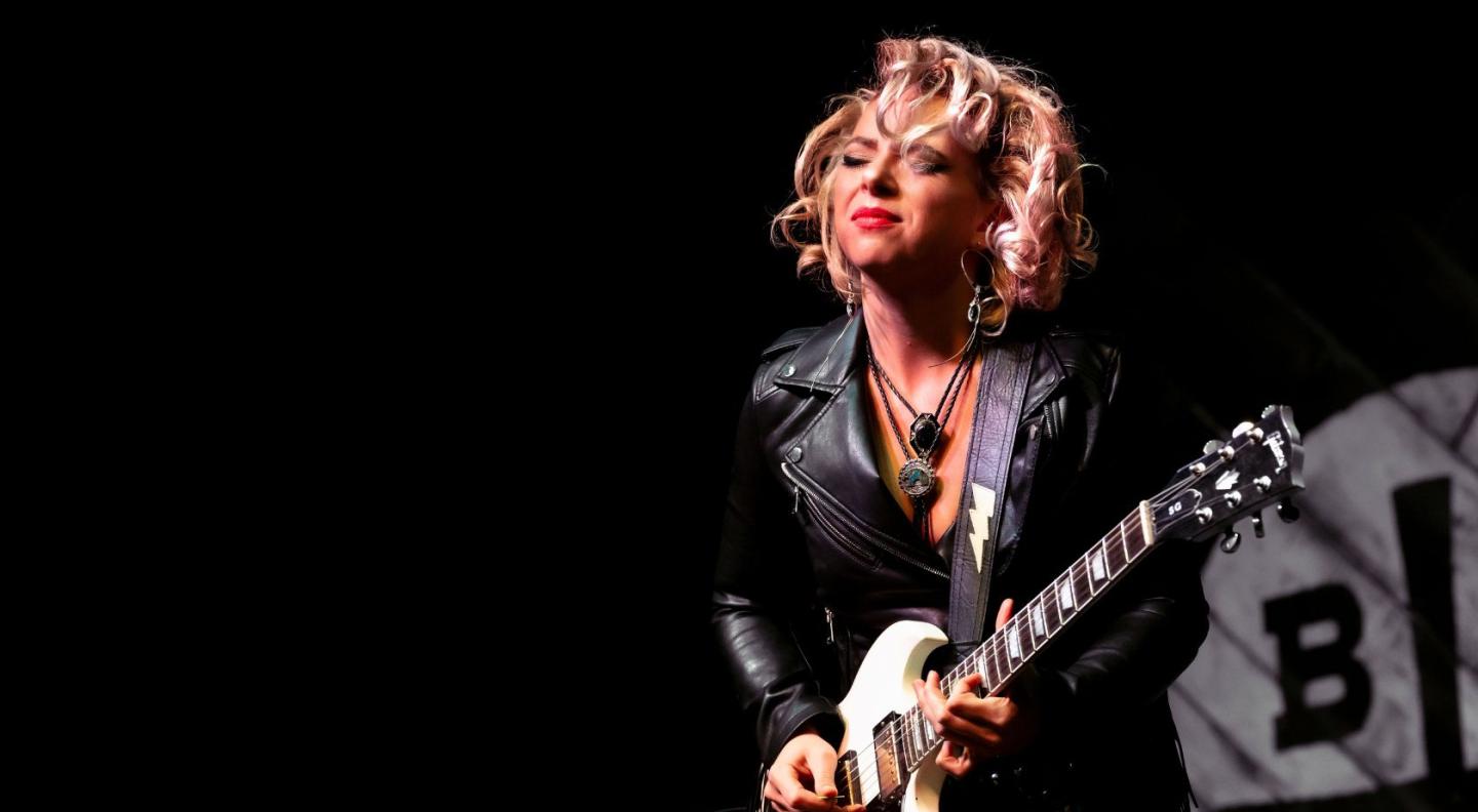 Samantha Fish, a white woman with short blonde hair, playing a white electric guitar in front of a black background.