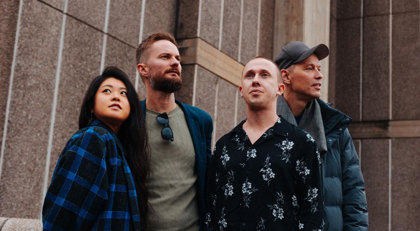 The four artists stand in front of a concrete wall, looking in different directions