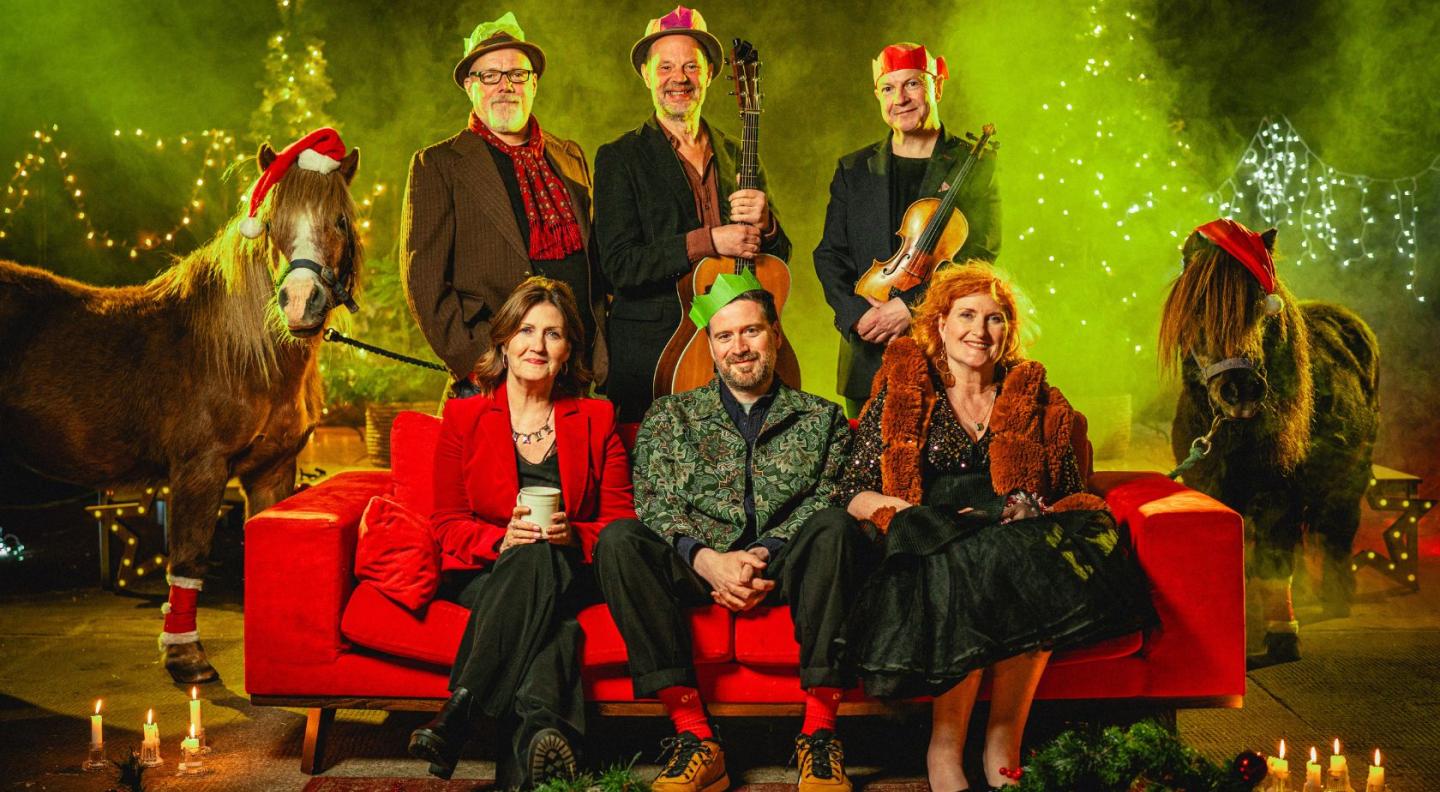 Musicians Eddi Reader, Karen Matheson, John McCusker, Kris Drever, Ian Carr and Kevin McGuire sitting on a sofa with two miniature ponies in santa hats