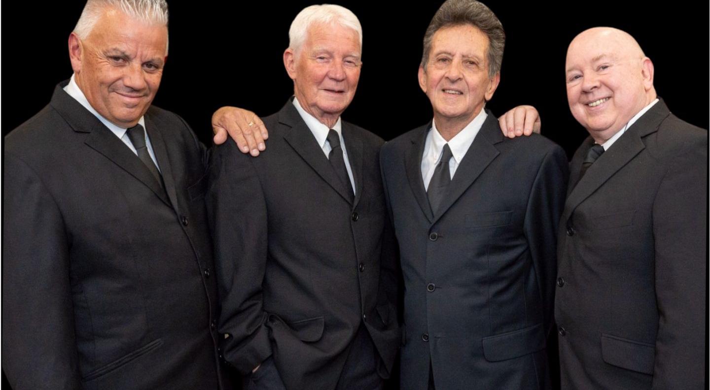 Four men in black suits stand against a black background