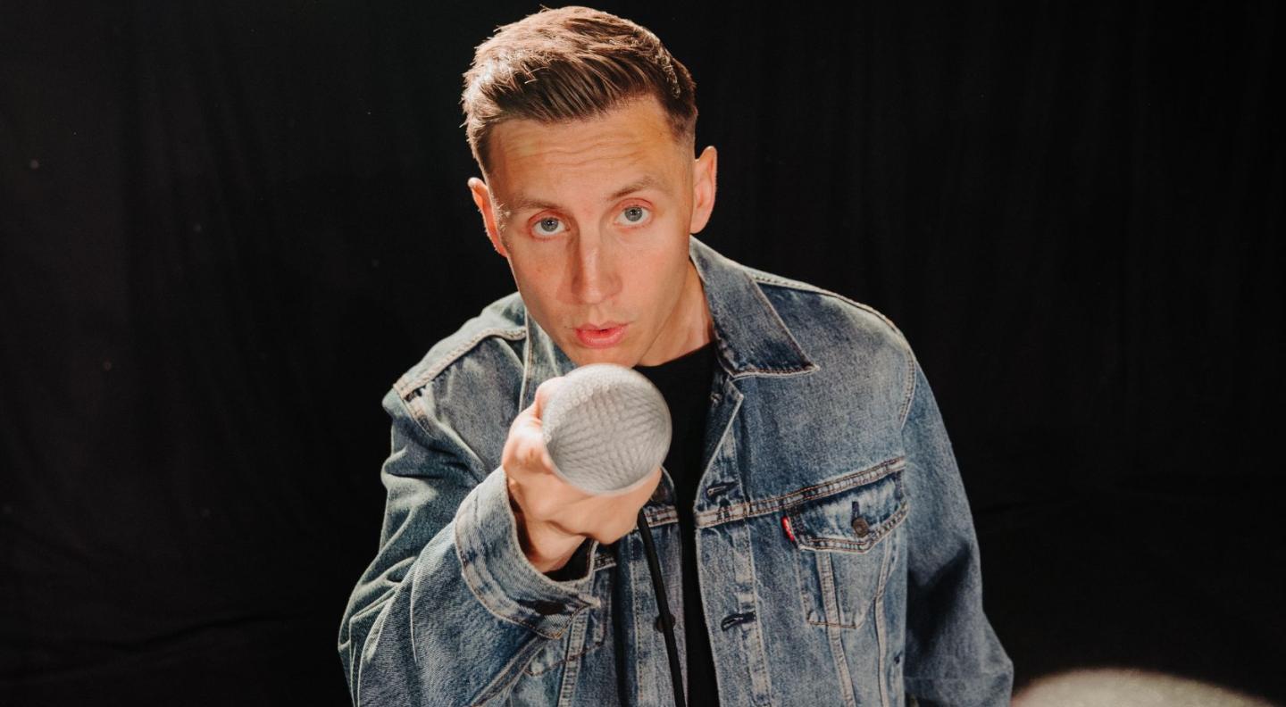 Shane Todd, wearing a denim jacket, looking directly into the camera, holding a microphone out towards it