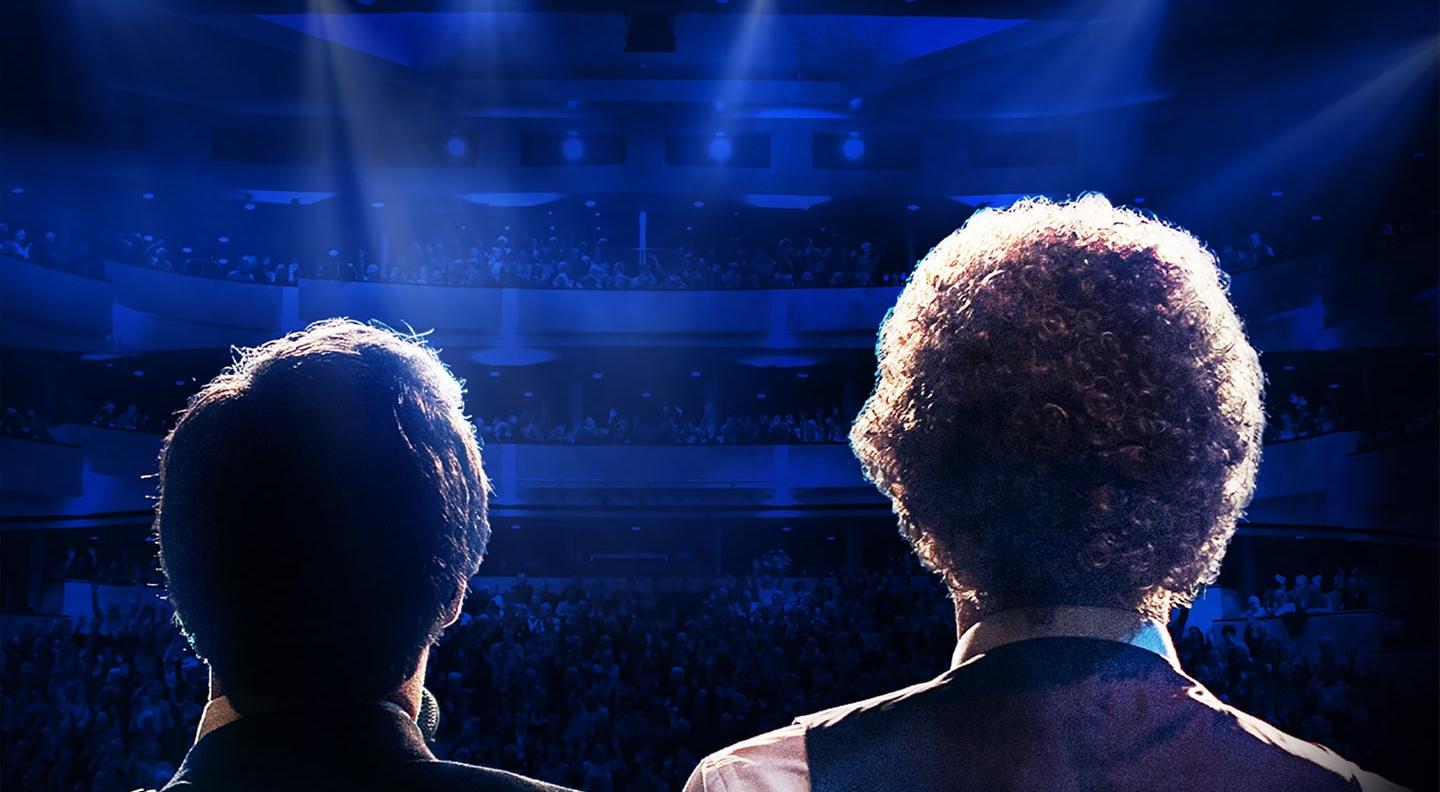 The back of two male performers' heads as they look out towards a lit auditorium