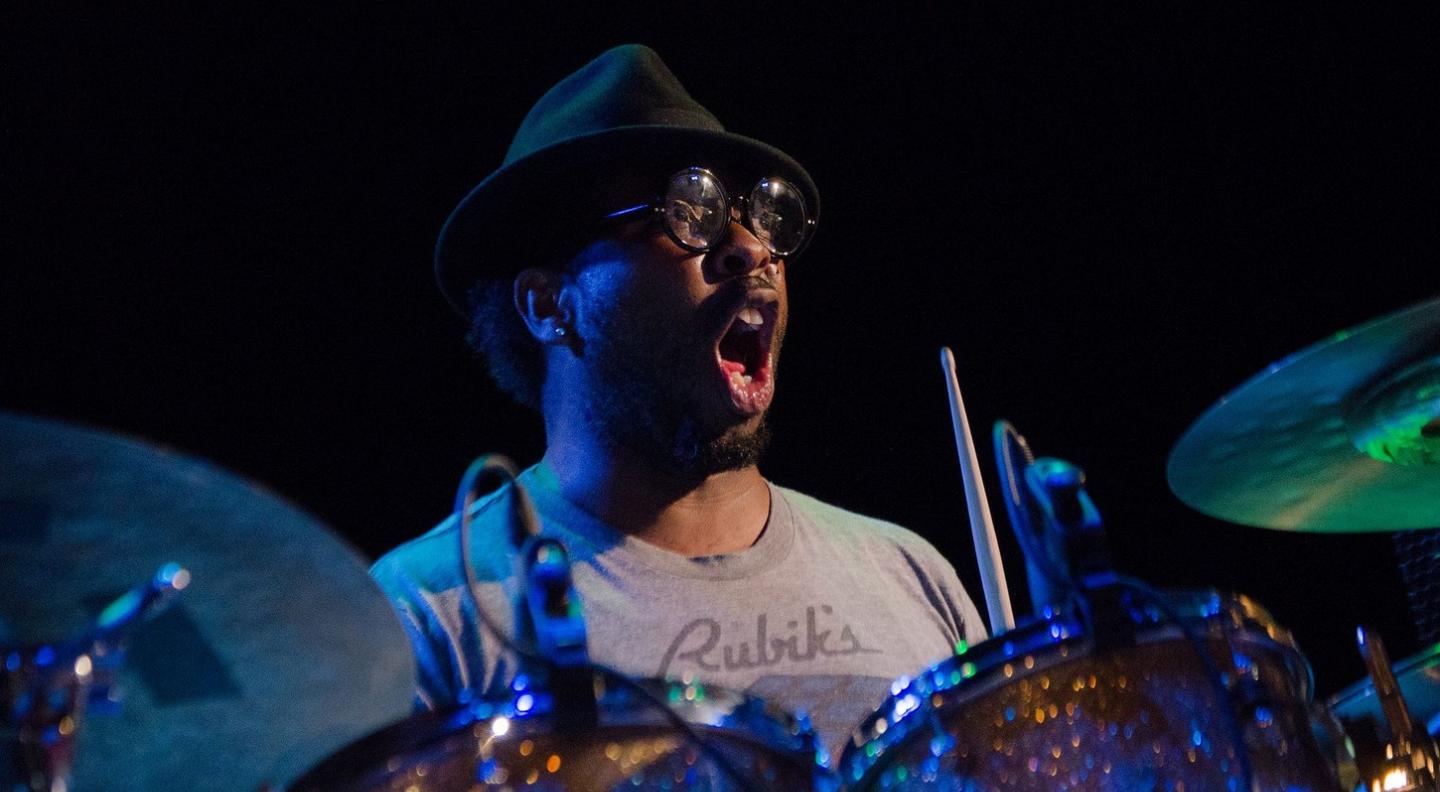A black man wearing a hat and glasses sits behind a drum kit holding the sticks in each hand with an open-mouthed surprised look on his face