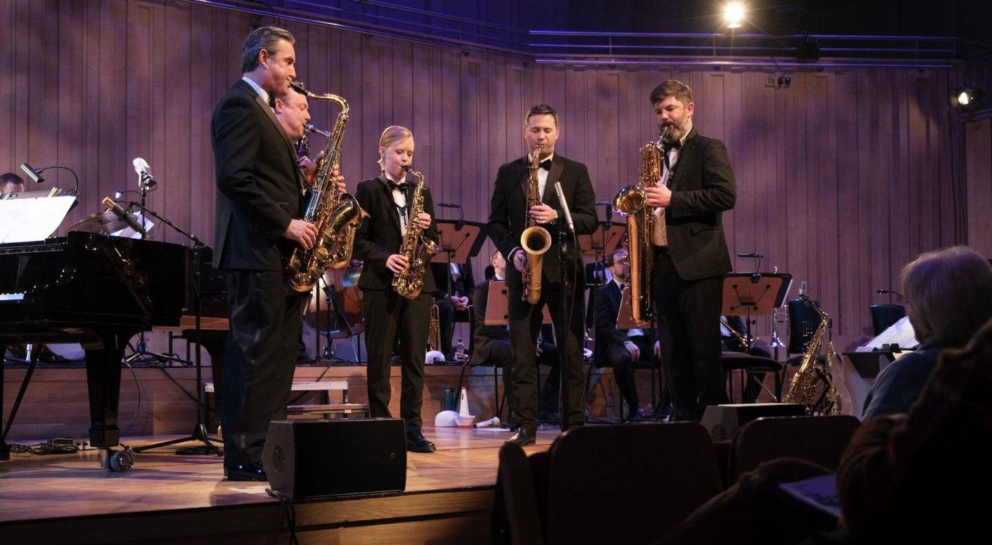 Five musicians stand on stage playing jazz instruments. They wear black suits and the stage is surrounded by  purple curtain