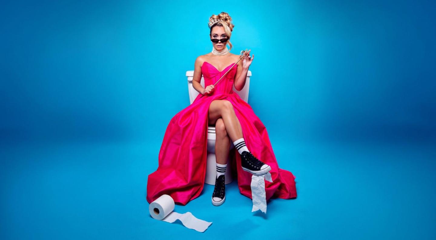 Sophie McCartney wearing a pink dress, a tiara and black sunglasses, sitting on a white toilet, against a blue background 