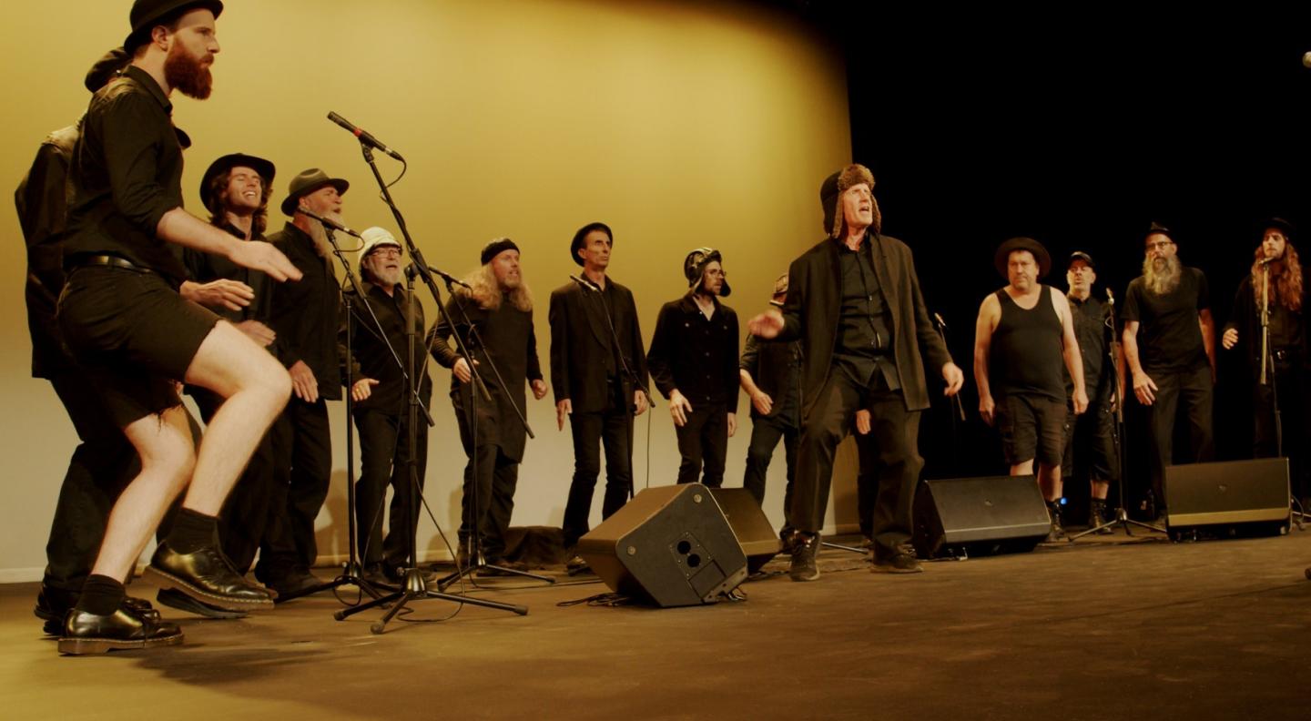 The Spooky Men's Chorale perform on stage in a line up with a yellowy light behind them
