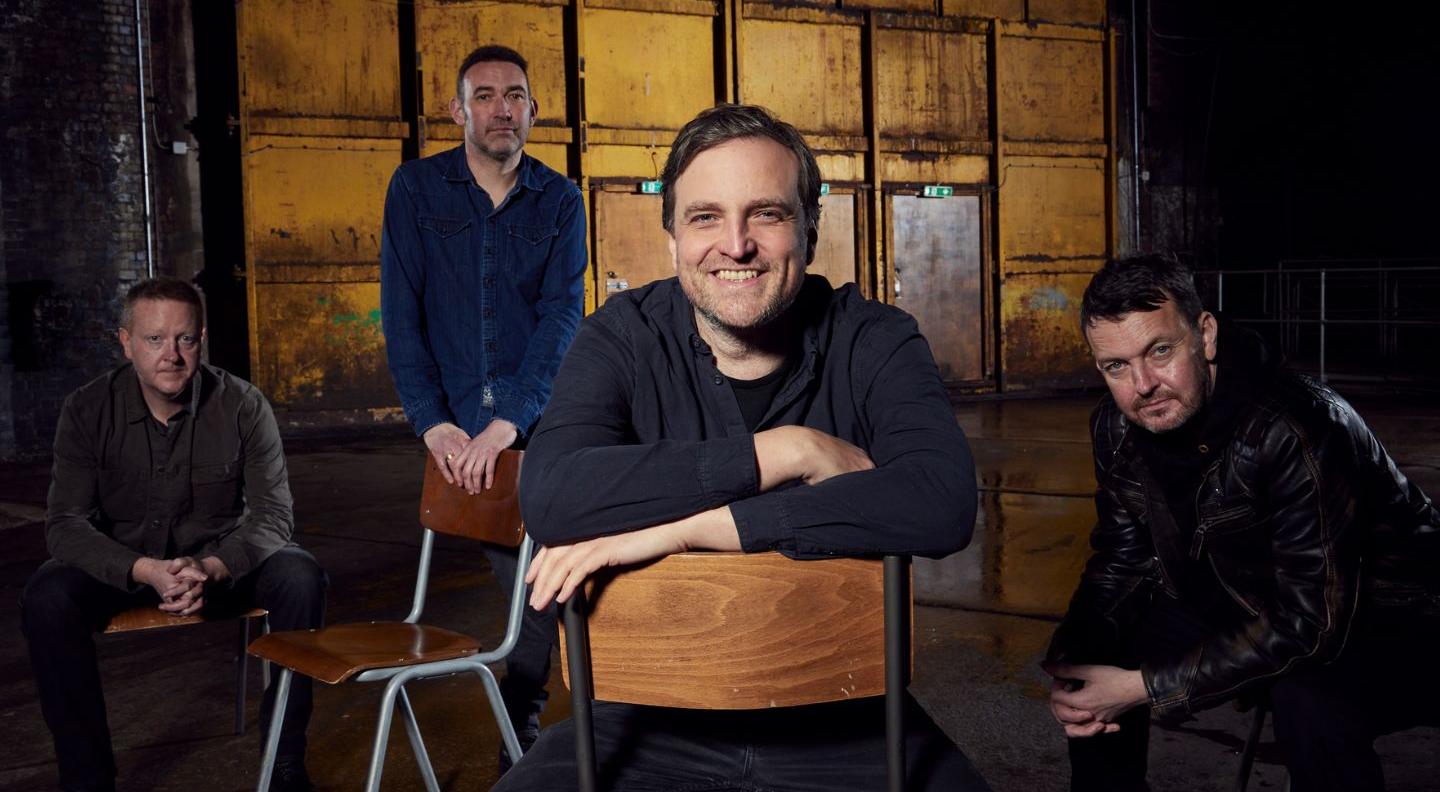 An image of the band starsailor sitting on chairs against a mustard yellow metal wall. The band are four white men with short hair in various colours. They all wear dark coloured clothing