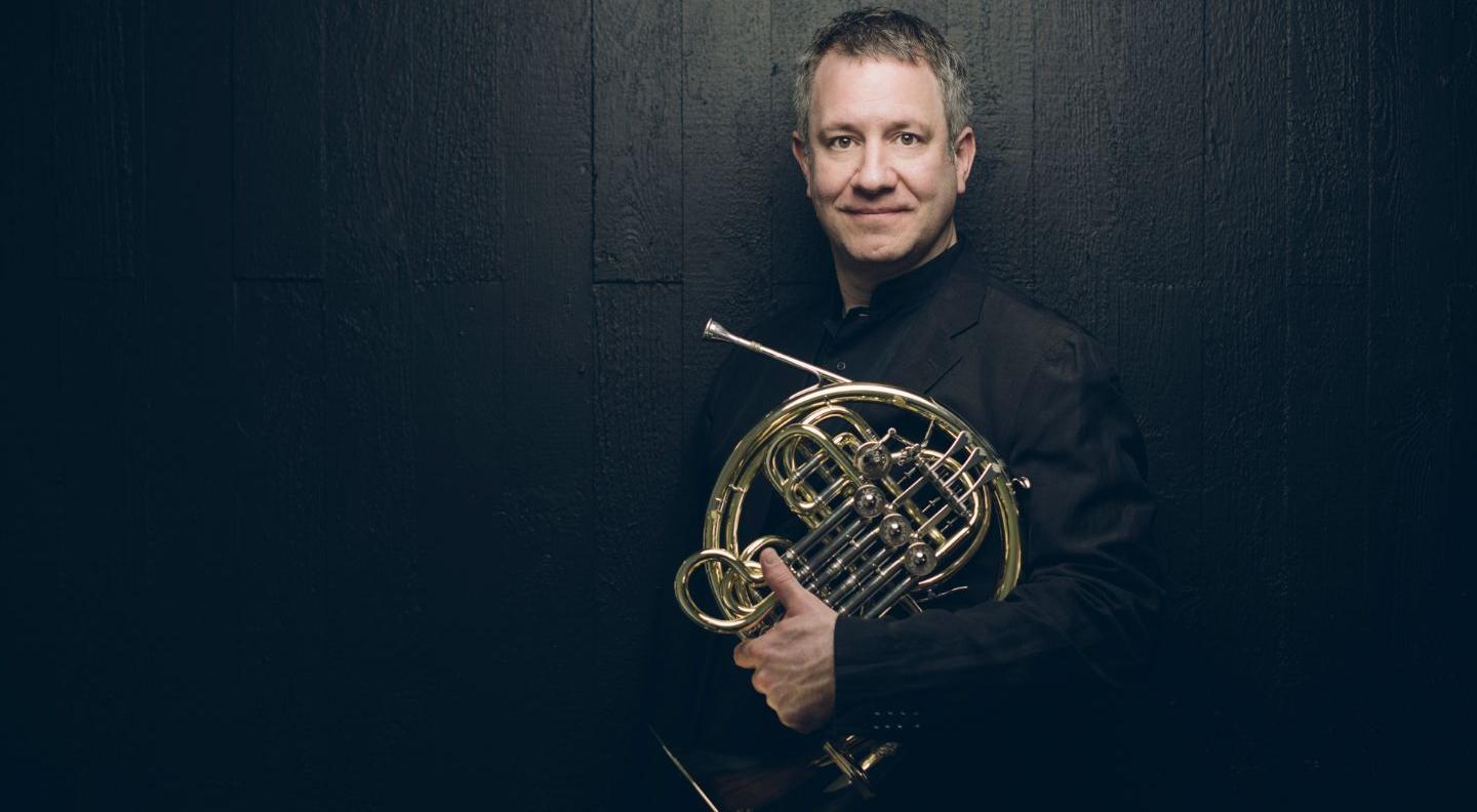 Stefan stands agains a dark wall, holding a french horn