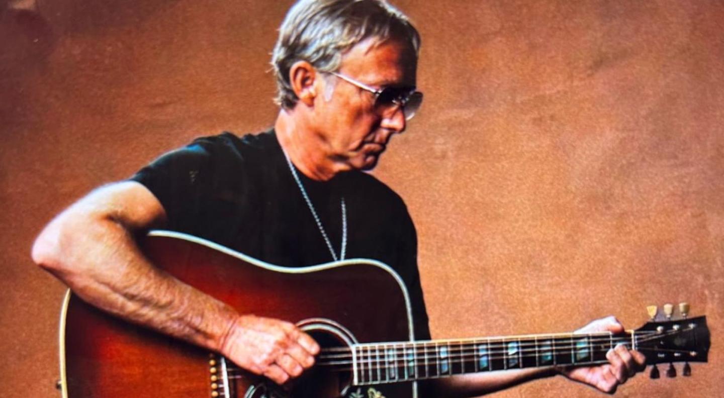 Steve Cradock playing the guitar sitting in front of a brown background