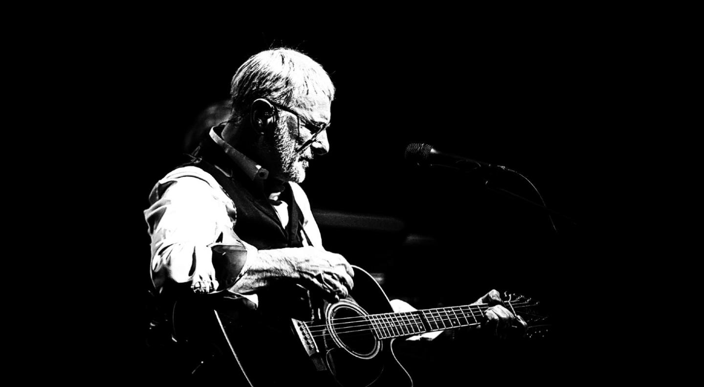 Steve Harley sits on stage playing guitar