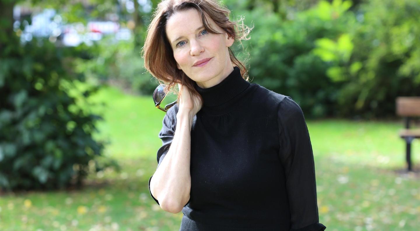 Susie Dent, dressed in black stands in a garden surrounded by green lawns and trees. Her head is cocked to her right and she has her bare right arm up holding it