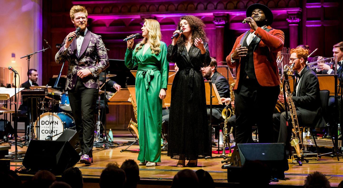 Four members of the band stand on stage in a line, all holiding microphones