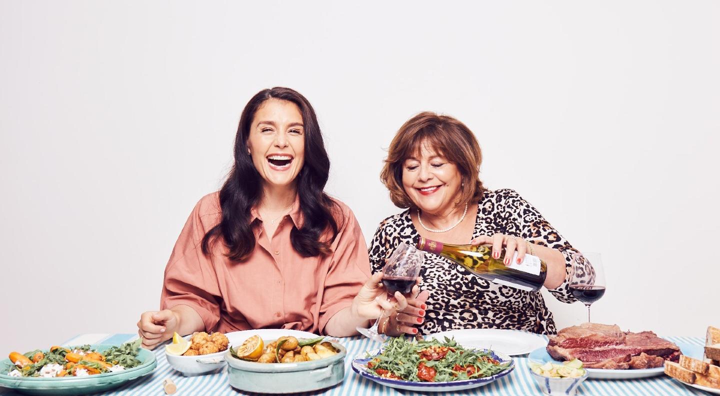 Jessie and Lennie Ware sit behind a table laden with delicious looking food. They're both laughing and Lennie is filling Jessie's glass with red wine