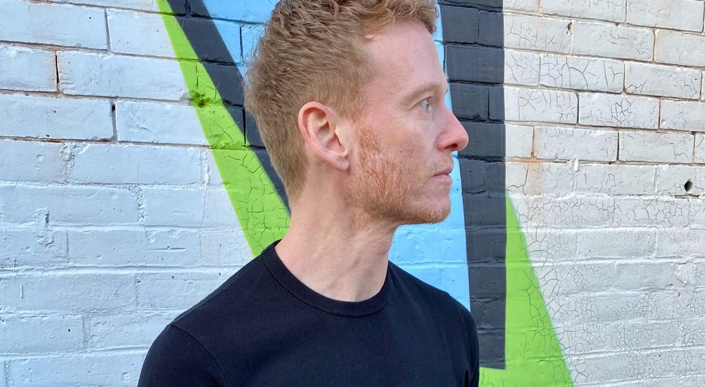 A mid shot of the artist Teddy Thompson, a white red headed man, looks off camera. He stands against a painted brick wall.